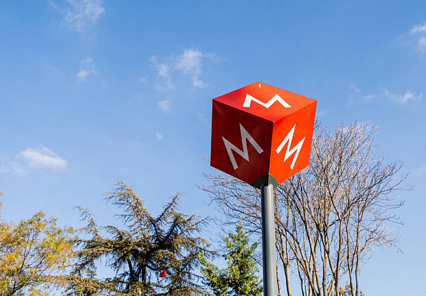 A red cube-shaped metro sign featuring the white “M” logo stands prominently against a bright blue sky, surrounded by autumn trees and natural daylight. This editorial photograph documents the exterior signage marking the entrance of the Ankara Metro system, highlighting public transportation infrastructure, urban mobility, and daily city life captured in a clear and detailed outdoor setting in Ankara, Turkey
