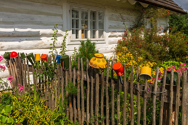 A row of colorful enamel mugs and utensils decorates an old wooden fence beside a blooming garden. The scene captures charming countryside aesthetics, handmade details and cozy homestead atmosphere.
