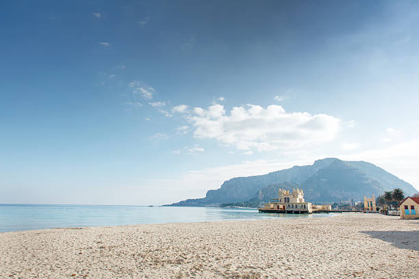 A tranquil coastal scene with turquoise water, soft sandy shore, and distant mountains, highlighted by a small historic pier building along the calm shoreline.
