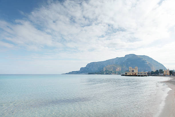 A tranquil coastal scene with turquoise water, soft sandy shore, and distant mountains, highlighted by a small historic pier building along the calm shoreline.