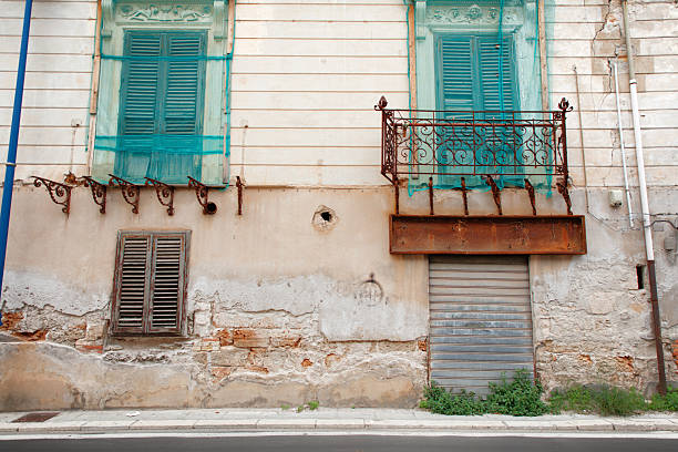 A weathered building facade features turquoise shutters, a decorative iron balcony, rusty railing, and a crumbling wall. The worn exterior evokes urban decay and historic charm along a quiet streetscape.