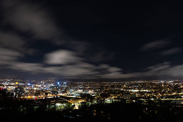 A high-resolution night view of Oslo, Norway, showcasing the illuminated city skyline under a dark sky filled with stars and drifting clouds. The long exposure captures smooth cloud movement and vibrant urban lights, highlighting the contrast between the calm night sky and the energetic city below. Ideal for themes related to travel, Scandinavia, urban life, night photography, and modern cityscapes.