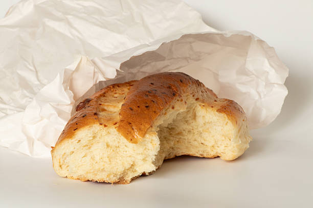 Fresh homemade bread, partially broken, is visible in a white paper bag against a light background, revealing the crust texture, soft crumb, and packaging. Baking concept
