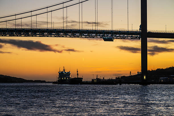 Cargo ship sailing under bridge at twilight with orange sky.