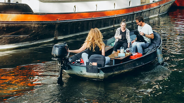 Editorial image taken on August 10, 2025, showing three individuals enjoying a casual outing on a motorized boat in an Amsterdam canal. One person steers from the rear, while the others sit around a small table with food and drinks. A small black dog is held by one of the passengers. The scene captures everyday leisure and social interaction in the Netherlands, with a larger vessel in the background and reflections on the water adding visual depth. This image documents urban life, canal culture, and the relaxed spirit of summer in Amsterdam.
