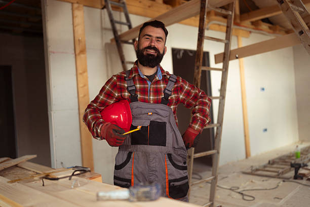 Male construction worker with beard wearing plaid shirt and grey overalls holding red safety helmet inside a wooden house under construction with tools and ladder in the background