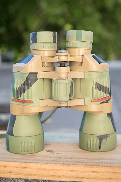 camouflaged binoculars stands upright on a wooden surface, with a blurred green outdoor background.