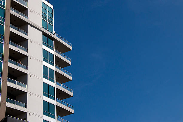 Woman on balcony of high-rise, San Diego Ca, November 3, 2025. A contemporary high-rise apartment building with glass balconies and reflective windows stands against a clear blue sky. The minimalist architecture and strong geometric lines create a clean, modern aesthetic. A woman stands on one of the balconies, adding a subtle human element without revealing identity.