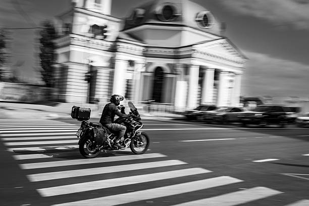 A solitary motorcyclist drives along a dusty rural road, framed by trees and fields in a remote countryside setting. The black and white composition emphasizes isolation, movement, and the timeless rhythm of rural life seen from a high vantage point.
