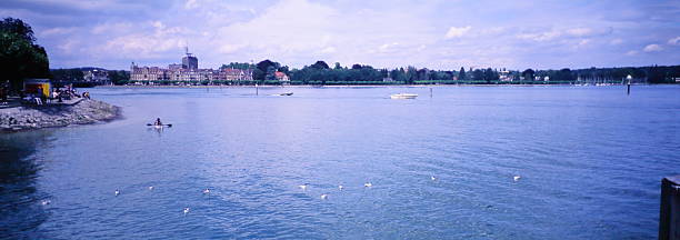 Tranquil panoramic scene across the shimmering waters of Lake Constance (Bodensee) toward the historic city of Konstanz, Germany, during the 2000s. A lone kayaker paddles near the foreground, while a small motorboat glides in the distance.