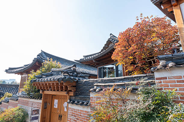Traditional Korean hanok house with tiled roofs and wooden doors surrounded by autumn trees with red and orange leaves. Historic architecture in a serene neighborhood setting. Korean culture and heritage.