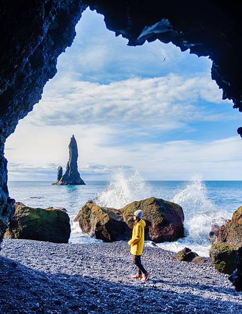 A traveler in a bright yellow jacket strolls along the pebbled shore of Reynisfjara Beach, framed by a caves opening in Iceland