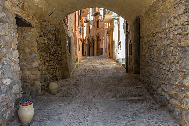 A timeless view down an arched stone passage to an historic street in Cefalu, Sicily, Italy. The medieval walls are rubble masonry - rough undressed fieldstones. Traditional uneven cobblestones, worn smooth over centuries. 2 large terracotta amphorae in front of a wooden door, 1 of which is being used as a ashtray. The street beyond showcases Sicilian vernacular architecture - stuccoed facades, balconies, wooden shutters, arched doorways