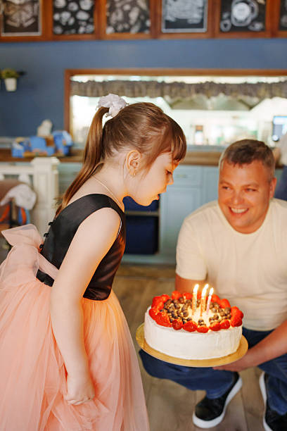 A young girl stands ready to blow out candles on a cake, while a smiling adult holds it. They are in a bright room decorated for a birthday party.