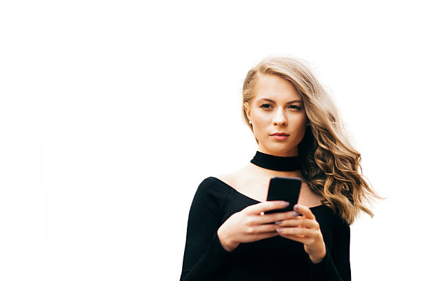 A young woman in a black outfit holds a smartphone with both hands while standing against a bright white background. Her focused expression and modern style create a clean, contemporary atmosphere. The minimal setting highlights themes of digital communication and connected lifestyle.