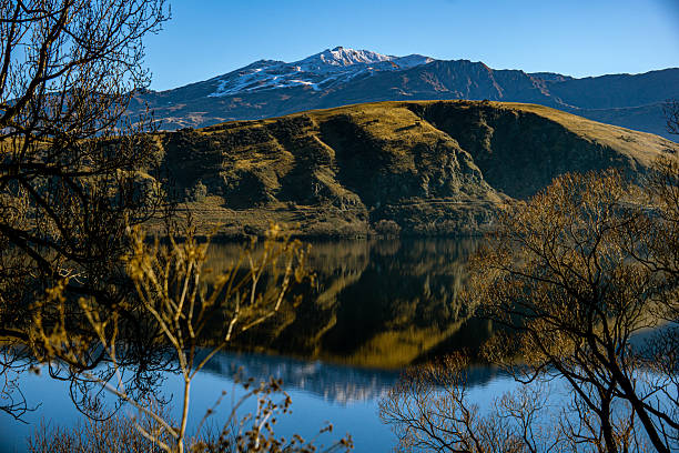 istock Winter's Tranquil Embrace: Snow-Capped Mountains Reflected in Calm Lake with Bare Branches in the Foreground Against a Bright Blue Sky 2246812335