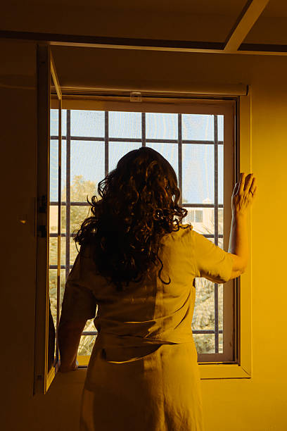 A woman stands looking outside of a window with bars in yellow dress with long hairs and feels locked up