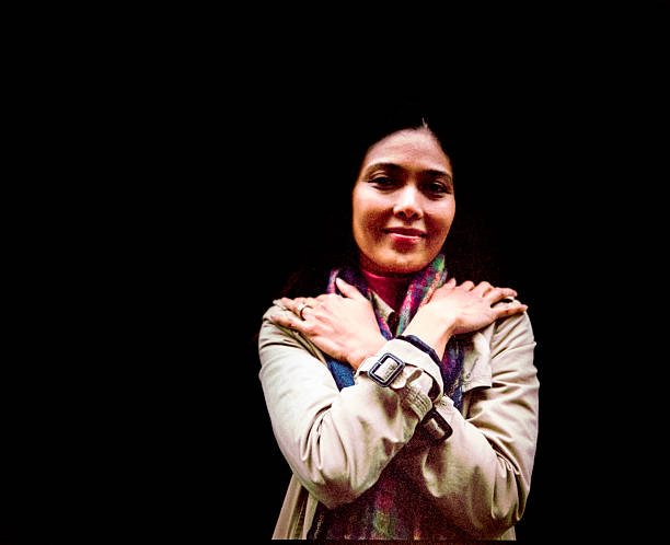 Filipino woman smiling confidently and embracing herself, wearing a beige trench coat and a colorful scarf, shot on analog film with visible grain, against a stark black background