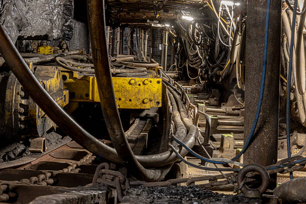 A dark, narrow mining tunnel stretches deep underground, illuminated only by faint artificial light. Dust, rock walls, and rails highlight the intense, labor-heavy atmosphere of active coal extraction