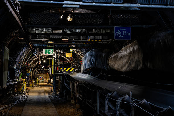 A dark, narrow mining tunnel stretches deep underground, illuminated only by faint artificial light. Dust, rock walls, and rails highlight the intense, labor-heavy atmosphere of active coal extraction