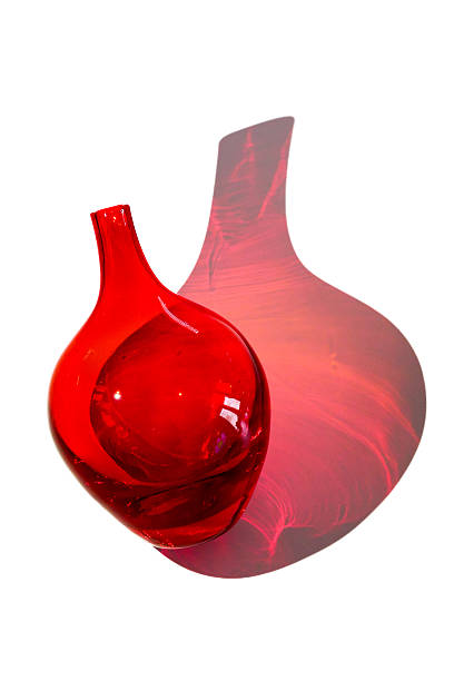 Decorative red glass bottle resting on a white background, illuminated by an intense light that passes through the glass and creates a wide, soft, diffused red shadow. The object has elegant and modern lines, ideal for interiors, minimalist design, furnishings, and artistic compositions. The image highlights transparencies, reflections, and plays of light.