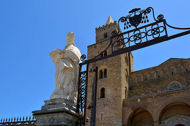 The ornate iron entrance gate to Cefalu Cathedral in Italy, featuring elegant symmetrical scrollwork, flanked by marble statues in classical flowing robes. 12th century Sicilian-Norman towers constructed of ashlar masonry on the upper levels with elegant arched bifore mullioned windows, crenellations at the top. 15th century Gothic portico comprising 3 pointed arches with marble columns. Interlaced blind arches in the central bay