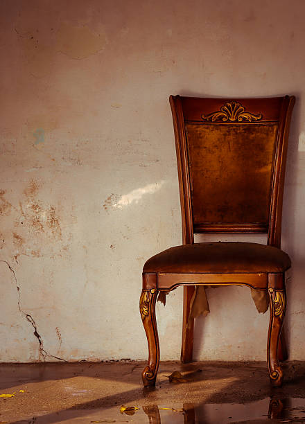 A moody, vertical photograph focusing on a single, ornate, antique wooden chair with upholstered velvet and worn leather. The chair sits directly against a pale, cracked, and heavily textured wall, which takes up most of the frame, providing ample copy space. A strong, warm light source from the right illuminates the chair and reflects off a puddle or wet spot on the floor in the foreground, creating a dramatic and nostalgic atmosphere. The image evokes feelings of abandonment, decay, forgotten elegance, and the passage of time.