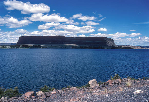 A tranquil lakeshore bordered by rocky cliffs and a mix of evergreen and bare trees. The calm water and textured rock formations create a natural, serene landscape that highlights the quiet beauty of untouched wilderness.
