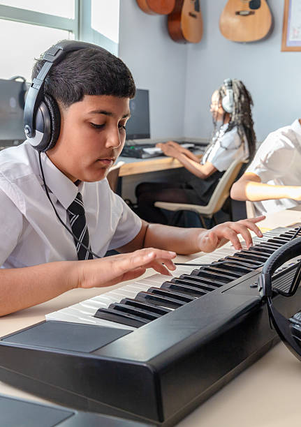 A close-up of a boy student in a school uniform seated at an electronic keyboard, concentrating on his music lesson. The scene captures a focused and dynamic environment for music education.
