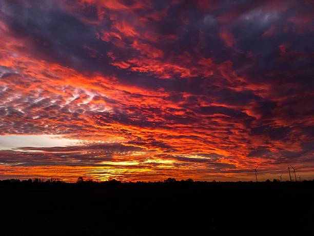 Vibrant red and orange clouds filling the sky during a dramatic sunset, creating a stunning backdrop over a silhouetted rural landscape with wind turbines on the horizon