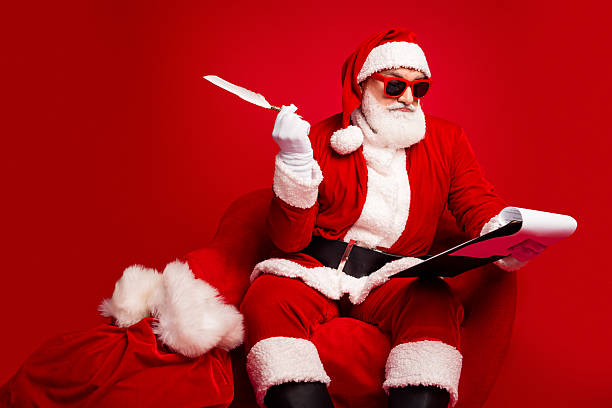 istock Santa in red suit with sunglass reads a script while holding a feather quill beside a gift sack in a bold red studio 2246870163