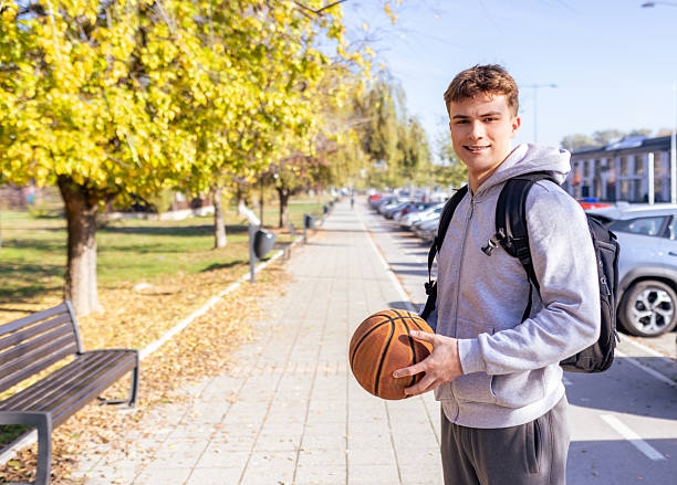 A smiling young male student stands on a sunlit sidewalk beside parked cars, wearing a gray hoodie and backpack while holding a basketball, capturing a casual, active moment of campus life outdoors.