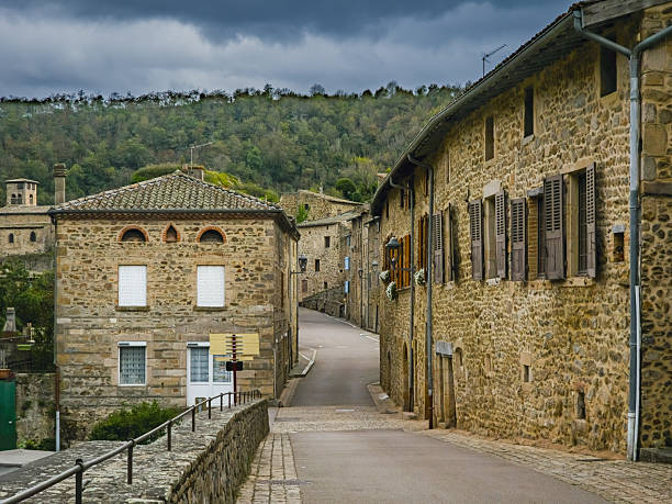 Editorial image of a scenic stone street in the village of Malleval, Loire department, France. Traditional French architecture with wooden shutters and empty cobbled street under a dramatic sky, surrounded by forested hills. Photographed on October 4, 2025.