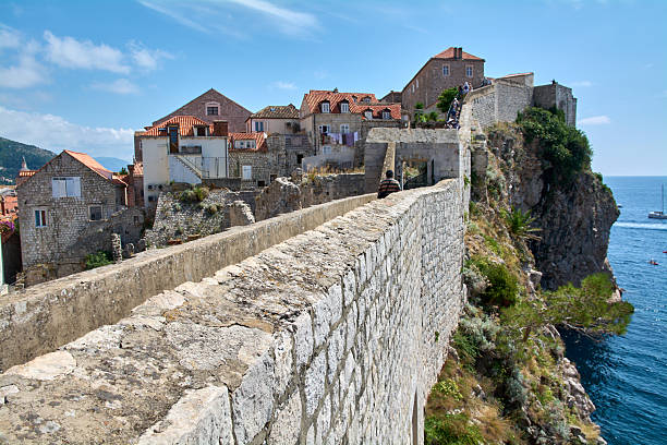 Dubrovnik. Croatia - November 15, 2025: Stone walls of Dubrovnik's old town on rocky cliffs above the blue Adriatic Sea. Tourists walk on the famous UNESCO landmark on a sunny summer day.