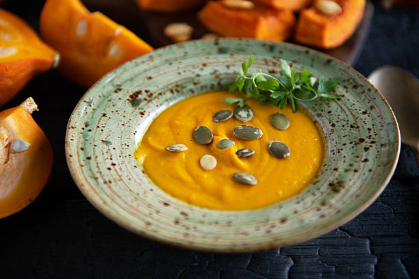 Overhead flat lay of pumpkin soup served in a rustic ceramic bowl with a spoon and fresh squash pieces on a dark wooden table. Cozy autumn cooking concept with warm seasonal colors.
