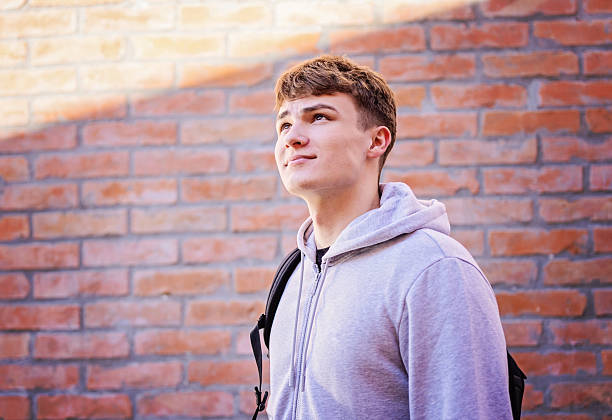 istock Young Man In Grey Hoodie With Backpack Gazes Upward Against Brick Wall 2246880603