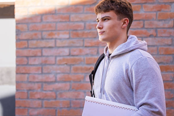 istock Young Man In Grey Hoodie With Backpack Holding Sketch Pad Beside Brick Wall 2246880606