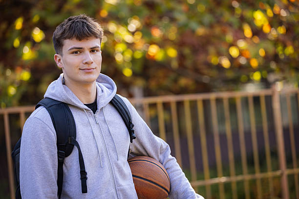 A teen student stands outdoors with a backpack and basketball, smiling softly. Autumn trees in the background create a warm, athletic vibe suitable for school, sports, or lifestyle imagery.
