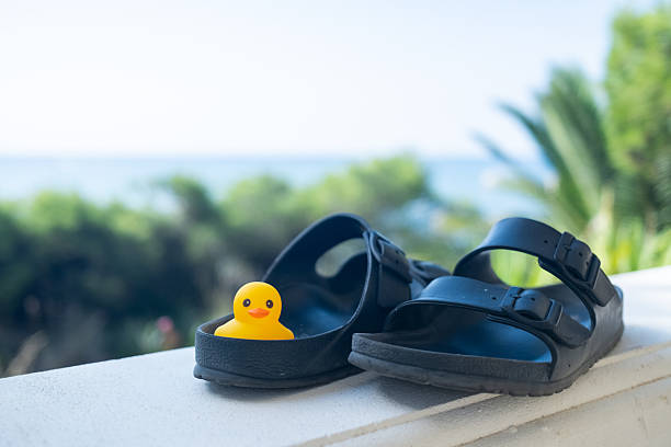 Yellow rubber duck toy resting inside a black sandal on a white wall with the ocean and green summer foliage in the soft focus background, symbolizing summer leisure and vacation fun