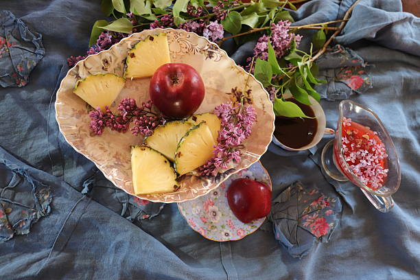 Healthy fruits in an antique vase—pineapple, apples. A cup of coffee on a light blue background among lilac flowers.