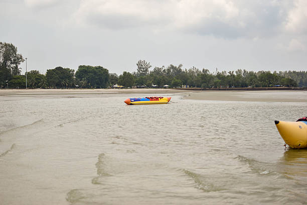 Inflatable boats rest in shallow beach water; yellow one with colorful seats stands out against cloudy sky and tree-lined shore.