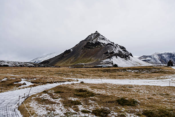 A landscape with a mountain partially covered in snow in a flat, dry grassland. The sky is cloudy.