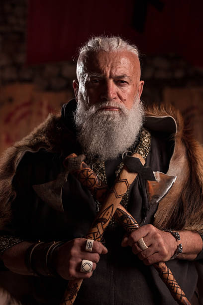 istock A formidable Viking chieftain with a striking white beard and braided hair, draped in a rich fur cloak, holds two crossed axes, gazing intensely into the camera in a dimly lit, rustic setting. 2246923966