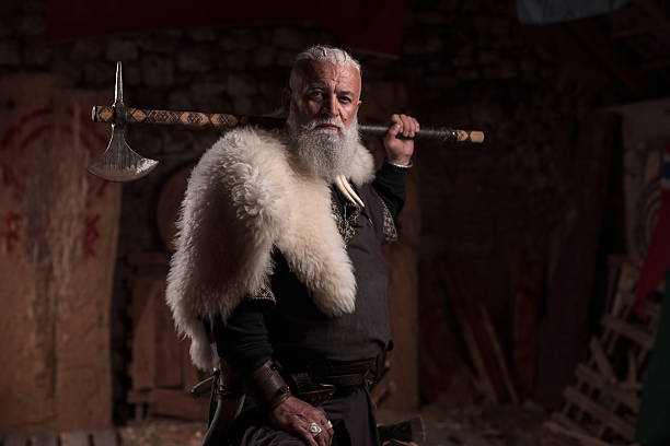 A resolute Viking chieftain, with a white beard and fur cloak, stands holding a formidable axe over his shoulder and a red and black round shield, embodying strength and ancient resolve.