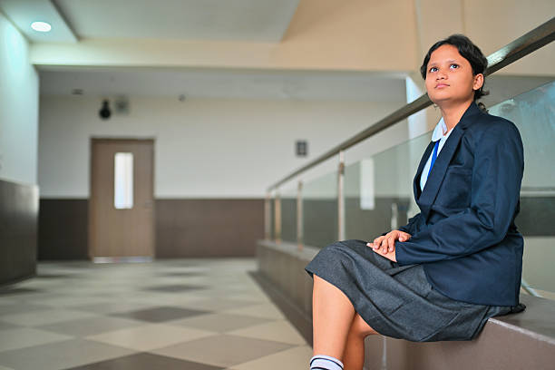 In a modern school hallway and soft lighting, a girl in a blazer and gray skirt sits on the ledge by the railing, hands resting on her lap and eyes lifted, creating a calm, reflective scene that highlights uniformed campus life.