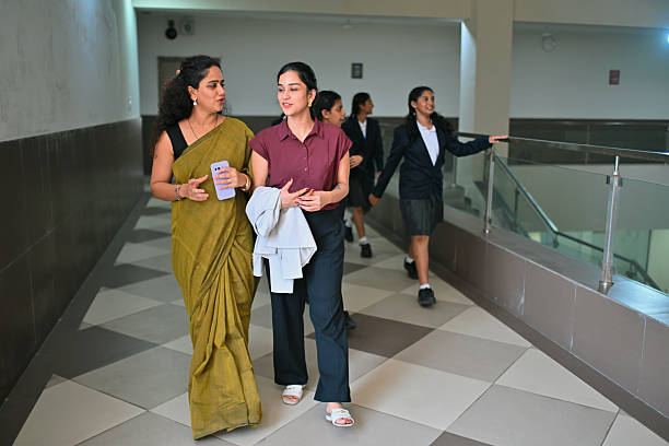 In a bright school hallway, a teacher in a sari and another in a blouse with a jacket draped over her arm stroll side by side, chatting warmly; behind them, uniformed students linger near the railing, adding a sense of campus life and collegial collaboration.