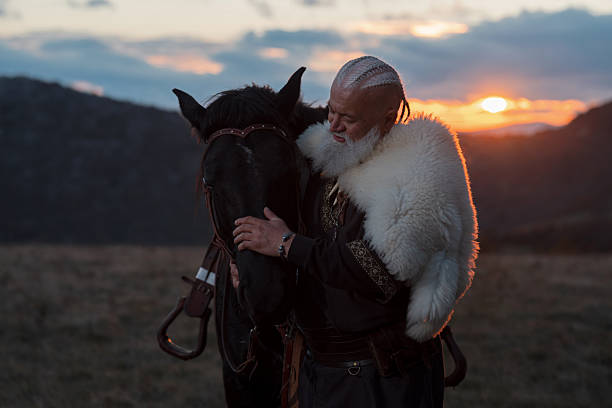 An elderly Viking warrior man with a white beard, dressed in traditional attire, standing confidently beside a horse at sunset, set against a scenic landscape.