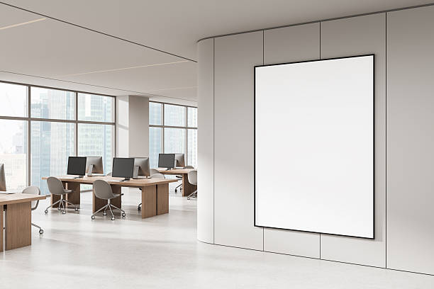 istock Blank poster mockup in modern open office interior with large windows, wooden desks and computer monitors in bright daylight setting. 3D Rendering 2246932059