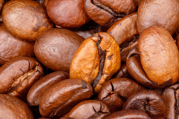 A tight, high-angle close-up shot showcases a pile of roasted coffee beans in various shades of brown, with visible cracks and textures.