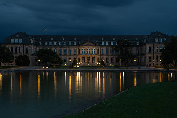 The New Palace in Stuttgart illuminated at night, reflected in a calm lake under dramatic evening clouds. A striking landmark and architectural nightscape.